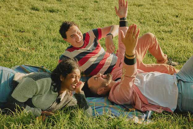 three young people having fun and laying on a picnic rug on the grass, image to promote supporting this cancer charity