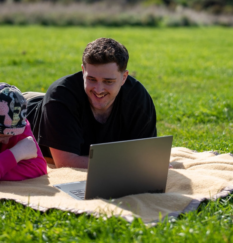 two young people laying on a picnic rug and accessing cancer counselling from canteen through their laptop